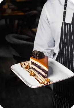 A waiter in striped apron is serving a gourmet slice of layered chocolate and caramel cake on a decorated plate in a restaurant.