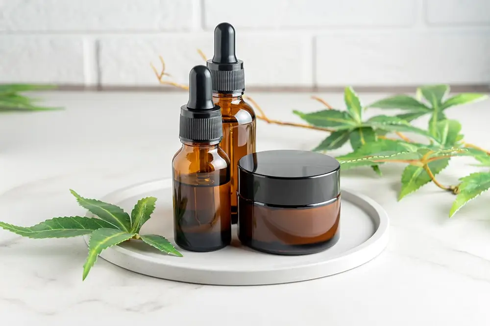 Brown, opaque cannabis oil bottles and a tub of cream surrounded by a cannabis plant on a marble counter.