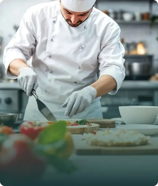 A chef in a white uniform preparing food in a commercial kitchen, illustrating culinary school practicals.