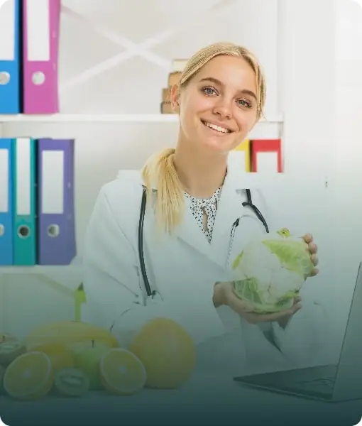 A smiling nutritionist in a white coat sitting at a desk with fresh fruits and vegetables.